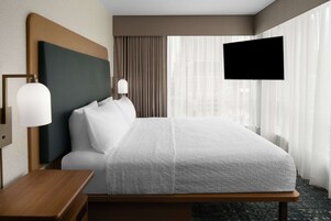 Habitación de hotel con una cama grande, una mesita de noche, un televisor de pantalla plana en la pared y una ventana con cortinas.