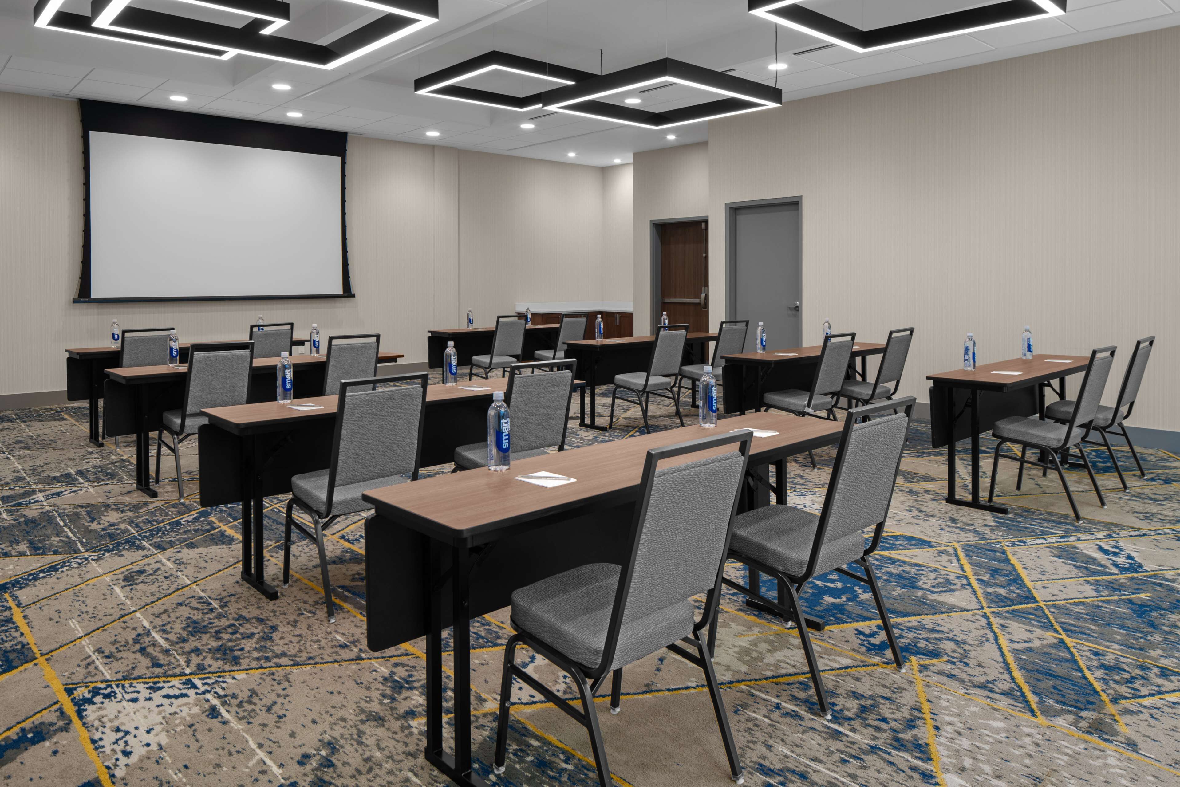 A conference room with multiple tables and chairs, a projector screen, and a carpeted floor.