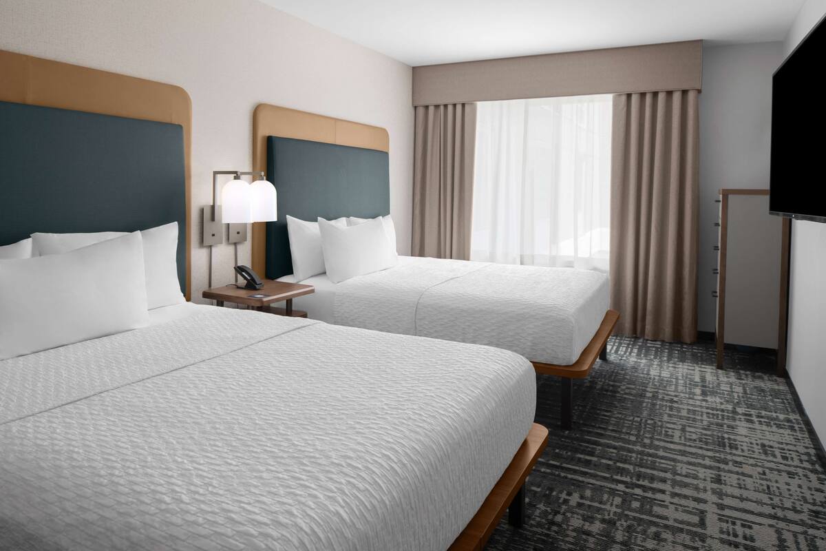 Ein Hotelzimmer mit zwei Betten, einem Schreibtisch, einer Lampe, einem Fernseher und einem Fenster mit Vorhängen.