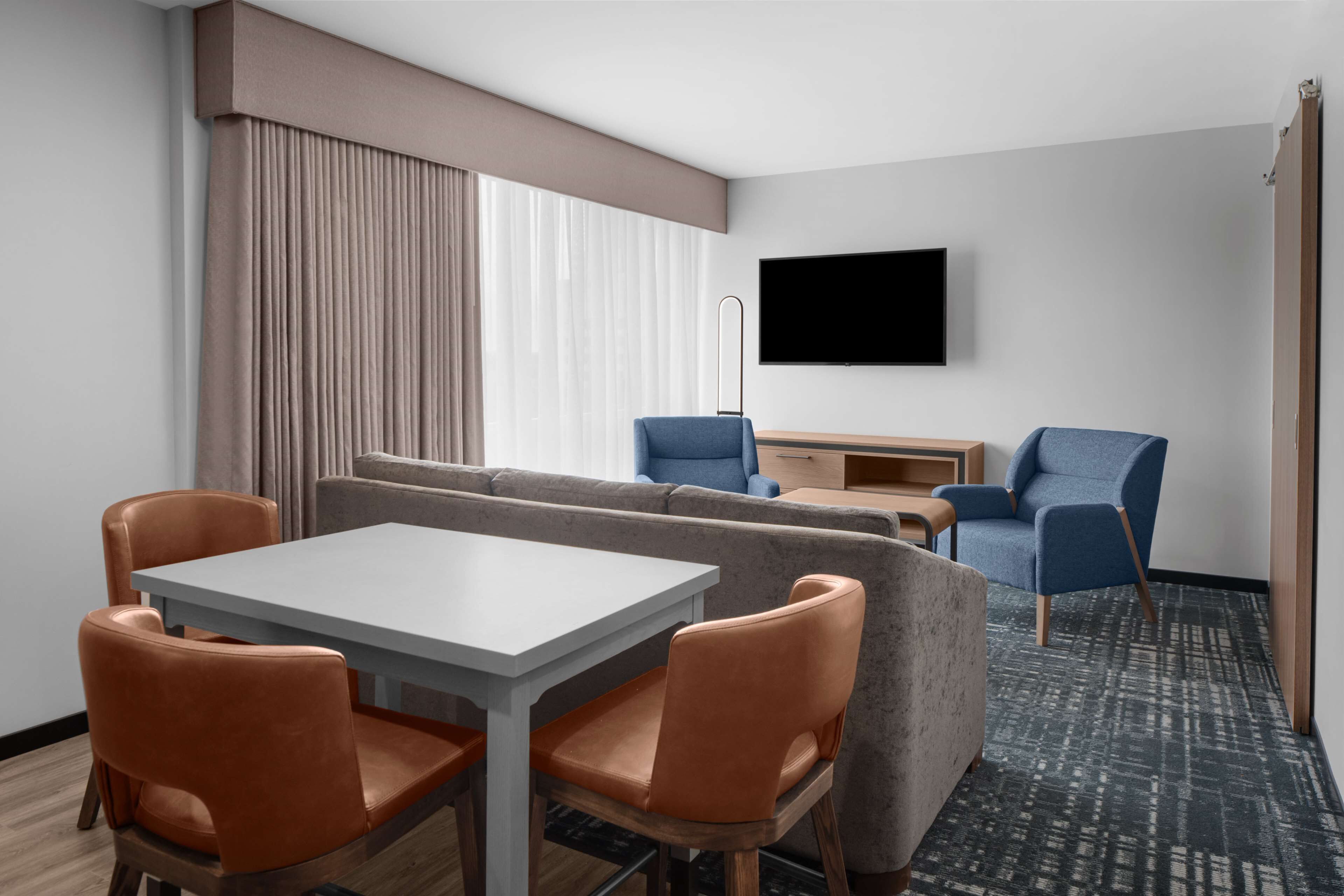 A hotel room with a dining table, two sofas, a TV, and a window with curtains.