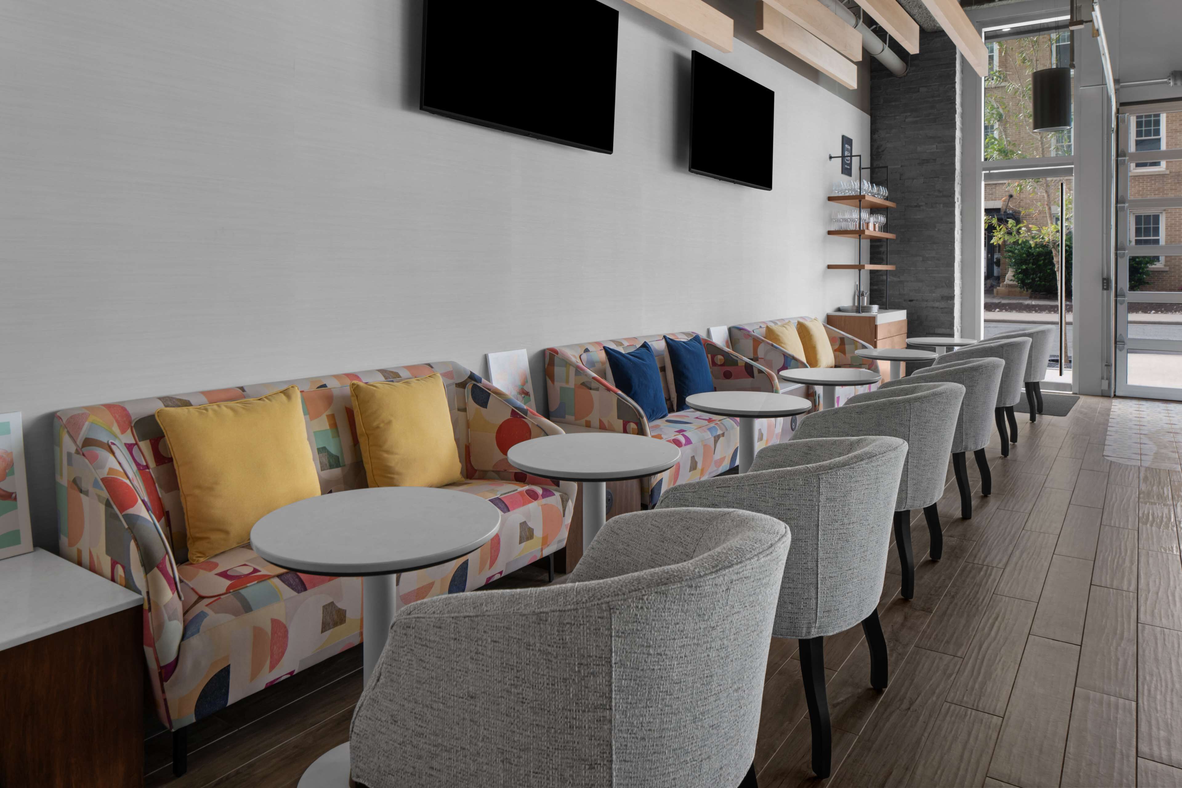 A modern lounge area with sofas and round tables, a flat-screen TV mounted on the wall, and a view of the outside through large windows.