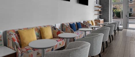 A modern lounge area with sofas and round tables, a flat-screen TV mounted on the wall, and a view of the outside through large windows.