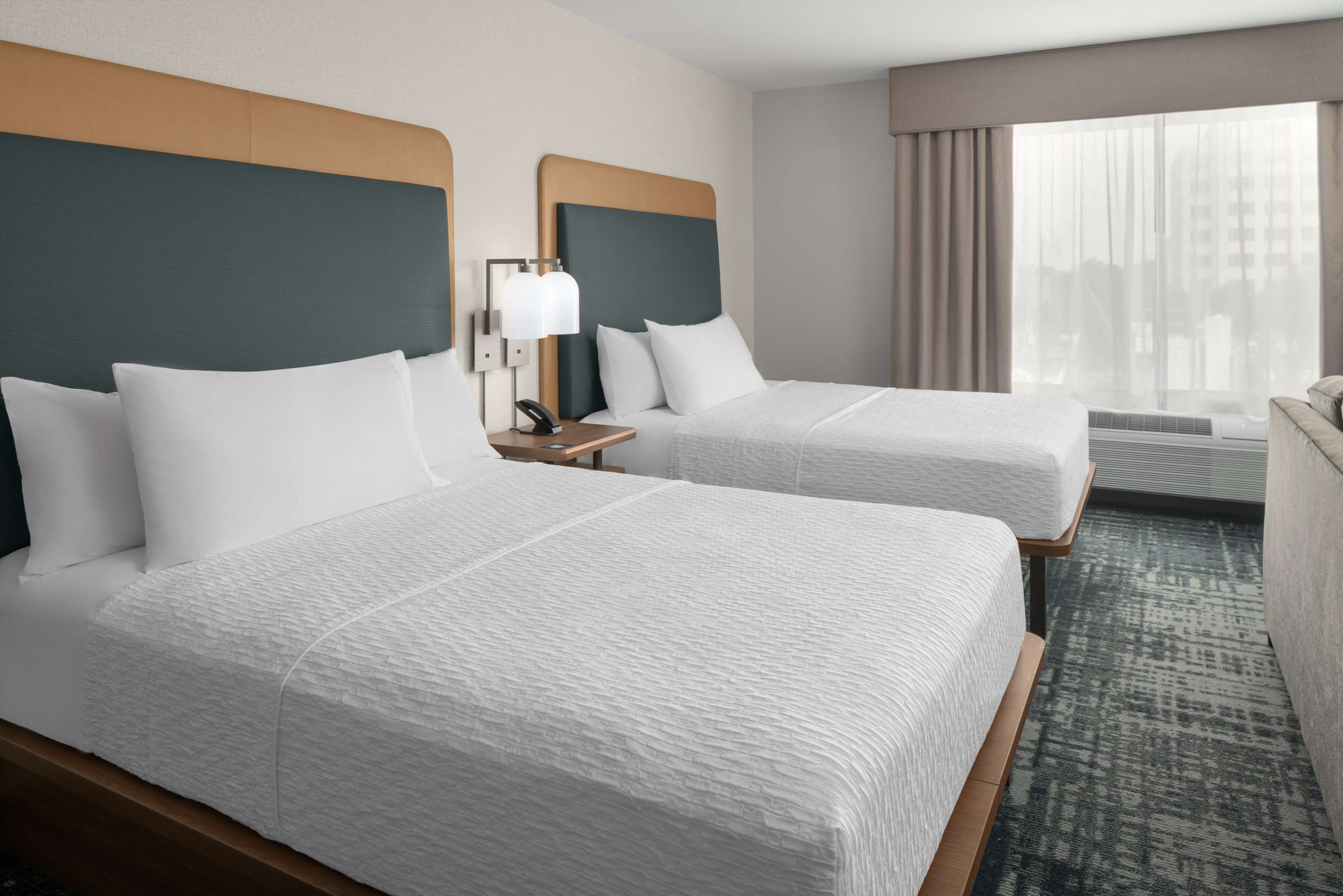 Quarto de hotel com duas camas, uma mesa de cabeceira com abajur, uma janela com cortinas e vista para edifícios.
