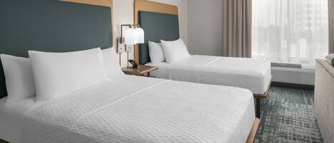 A hotel room with two beds, a nightstand with a lamp, a window with curtains, and a view of buildings.