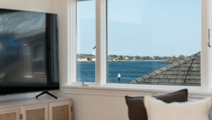 A room with a television on a stand, a window with a view of a body of water and buildings, and a cushioned seating area.