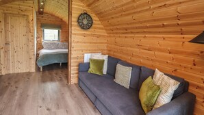 Ein gemütliches Cabin-Interieur mit Holzbalkendecke, einem grauen Sofa mit Kissen, einem Bett mit gemustertem Überwurf und einer Wanduhr.
