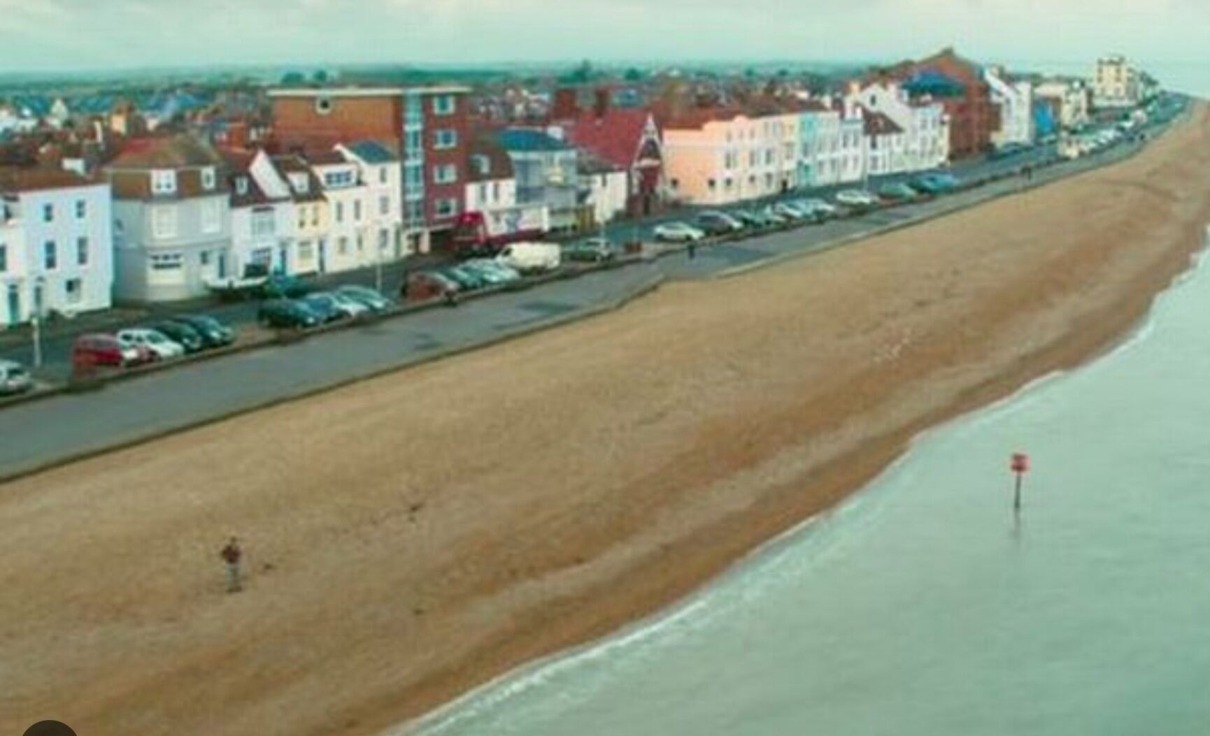 A coastal town with a long sandy beach, a row of colorful buildings, and parked cars along the shore.