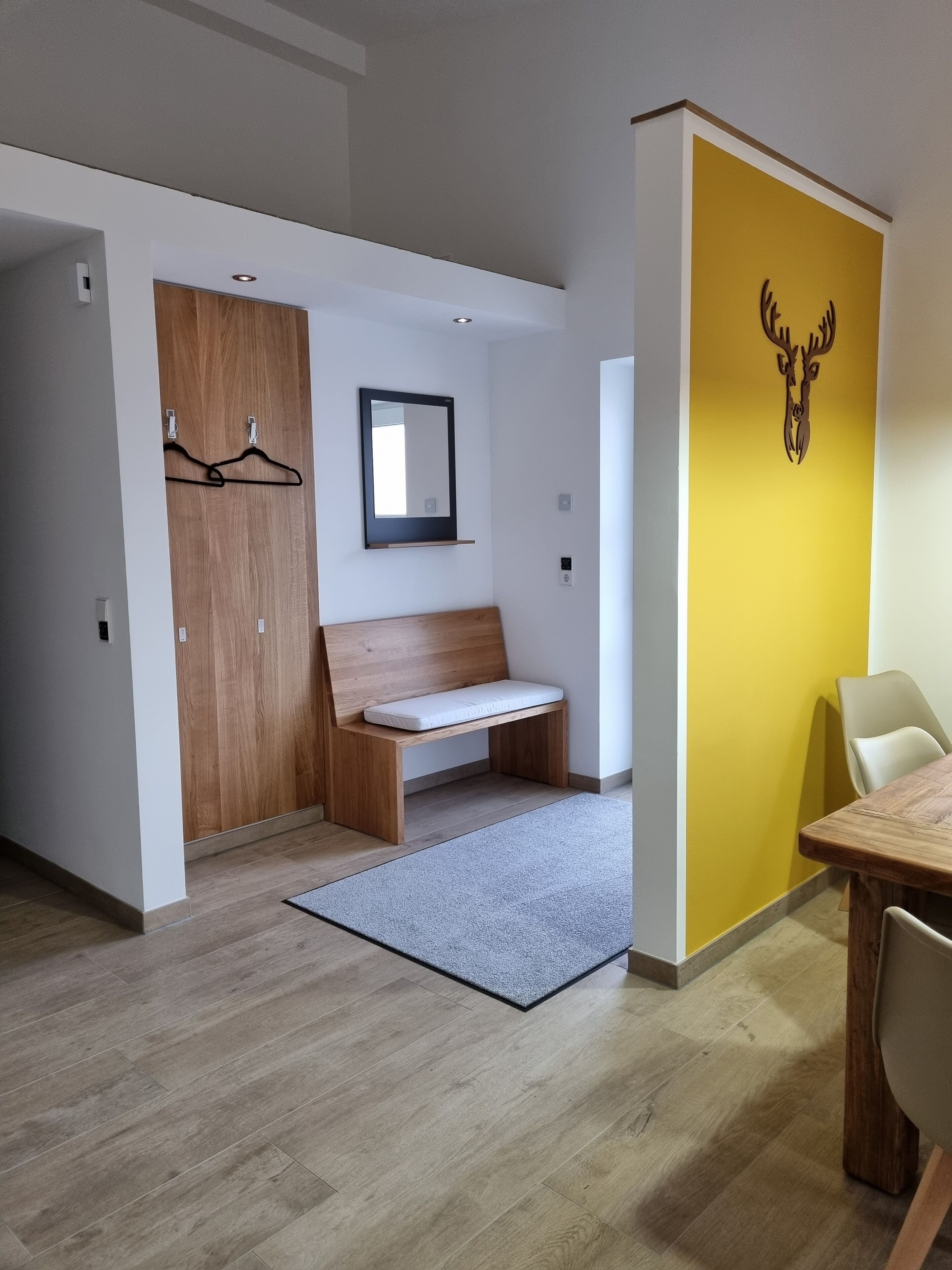 A modern room with a wooden bench, a small table with chairs, a mirror, and a wall-mounted deer head decoration.