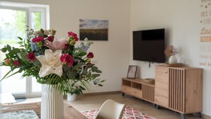 Un comedor con una mesa de madera, un jarrón blanco con flores, un televisor y un mueble de madera.
