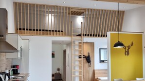 Una cocina moderna con estufa integrada, un comedor con mesa y sillas de madera, y un altillo con barandilla de madera.