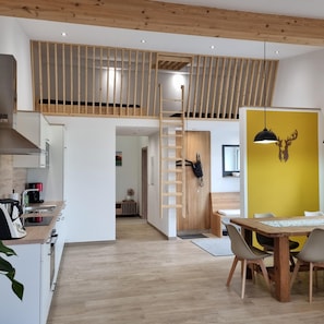 A modern kitchen with a built-in stove, a dining area with a wooden table and chairs, and a loft space with a wooden railing. - Aussiedlerhof in Alleinlage. Großzügige Ferienwohnung (Rot am See)
