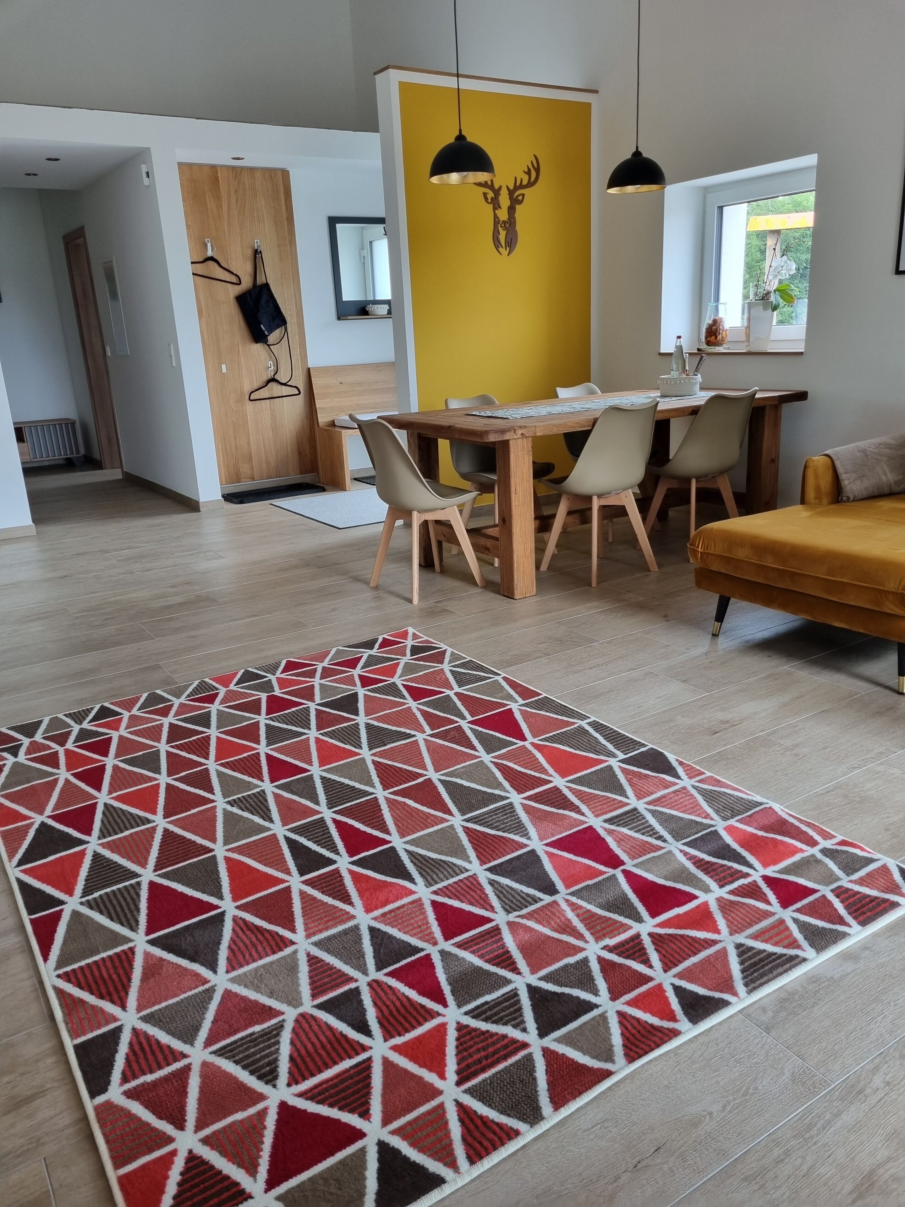 Ein modernes Wohnzimmer mit einem Holzesstisch, Stühlen, einer gelben Akzentwand und einem Teppich mit roten und grauen Mustern.