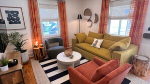 A cozy living room with a sofa, armchair, and a coffee table. There are two windows with curtains, a ceiling fan, and a wall-mounted lamp. - Millhouse in Downtown Jefferson (Jefferson)
