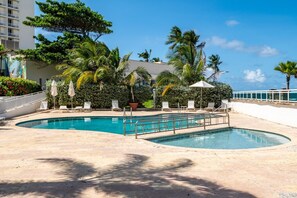 A pool area with lounge chairs and umbrellas, surrounded by palm trees and a building.