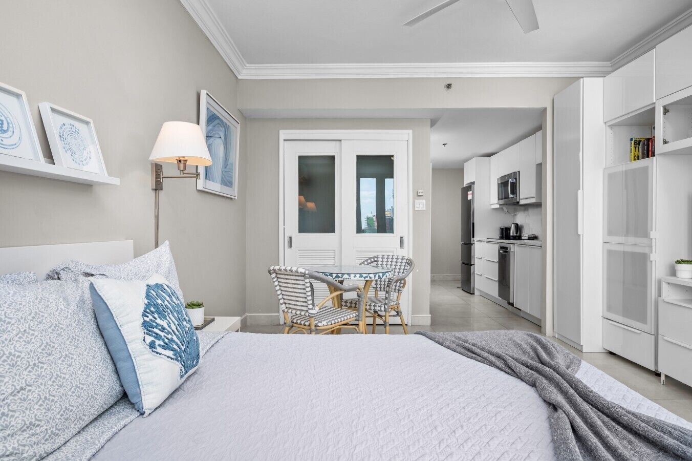 A bedroom with a bed, a bedside table, a lamp, a chair, a dining table, and a view into the kitchen.