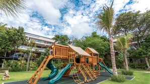 A wooden playground with slides and climbing structures, surrounded by lush greenery and a building in the background. - Awa 107 At AWA Residences - Playacar ll (Playa del Carmen)
