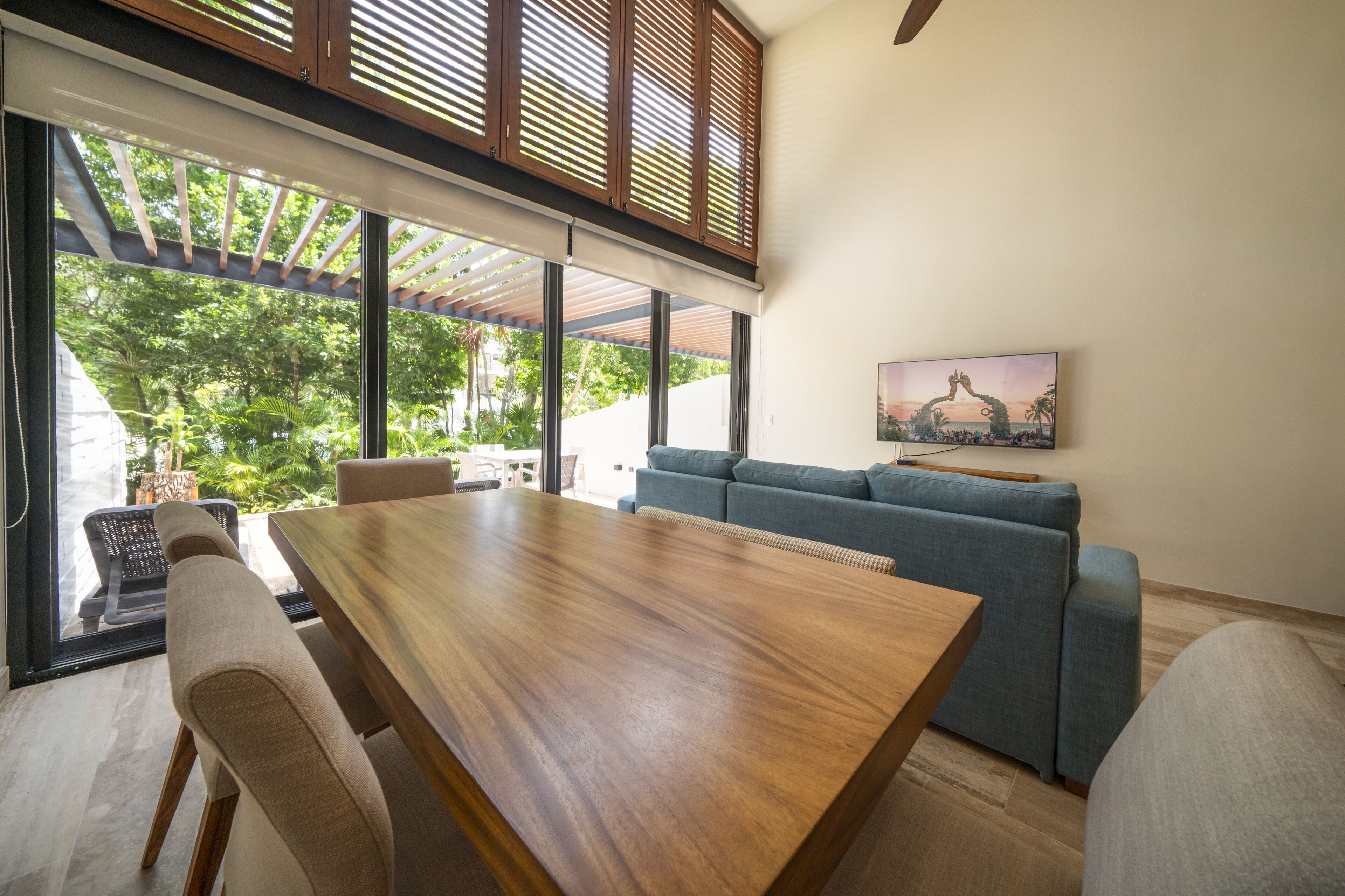 Una sala de estar moderna con una mesa de comedor de madera, un sofá azul y ventanas amplias que dan a vistas de follaje exterior.