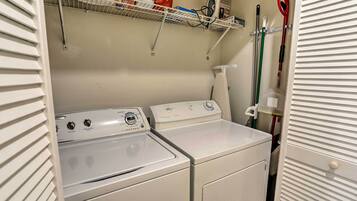 A laundry area with two white washing machines, a shelf with cleaning supplies, and a mop on the wall.