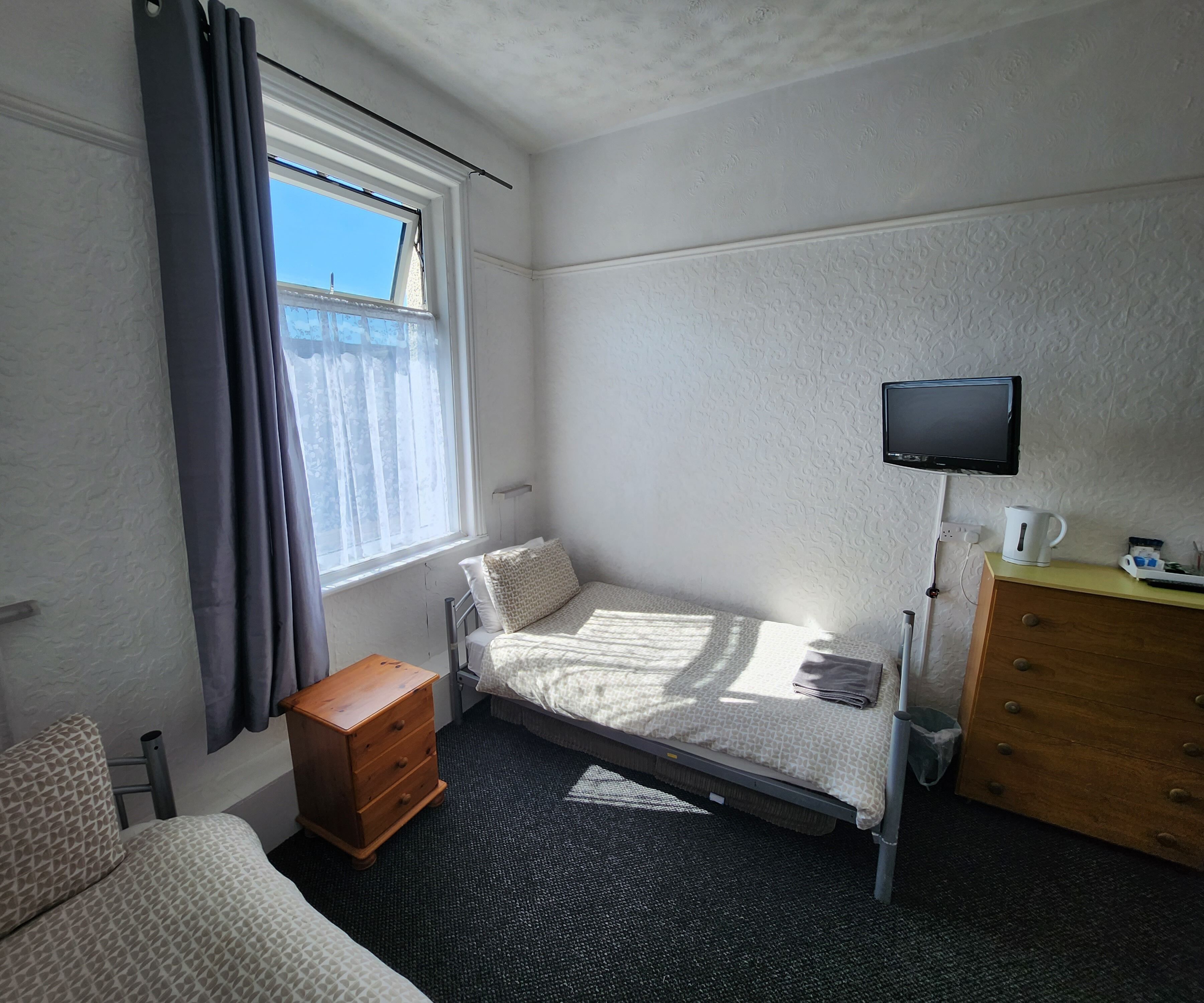 A small, well-lit room with a single bed, a wooden dresser, a television mounted on the wall, and a window with curtains.