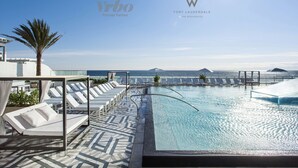 Ein Infinity-Pool auf dem Dach mit weißen Liegestühlen und einer Palme, mit Blick auf den Ozean.