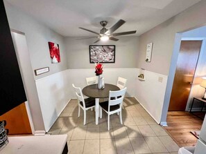 A dining area with a round table, white chairs, a ceiling fan, and wall-mounted artwork.
