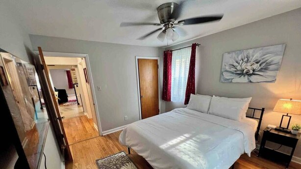 Ein Schlafzimmer mit Bett, Deckenventilator, Nachttisch, Lampe und einem Bild an der Wand.