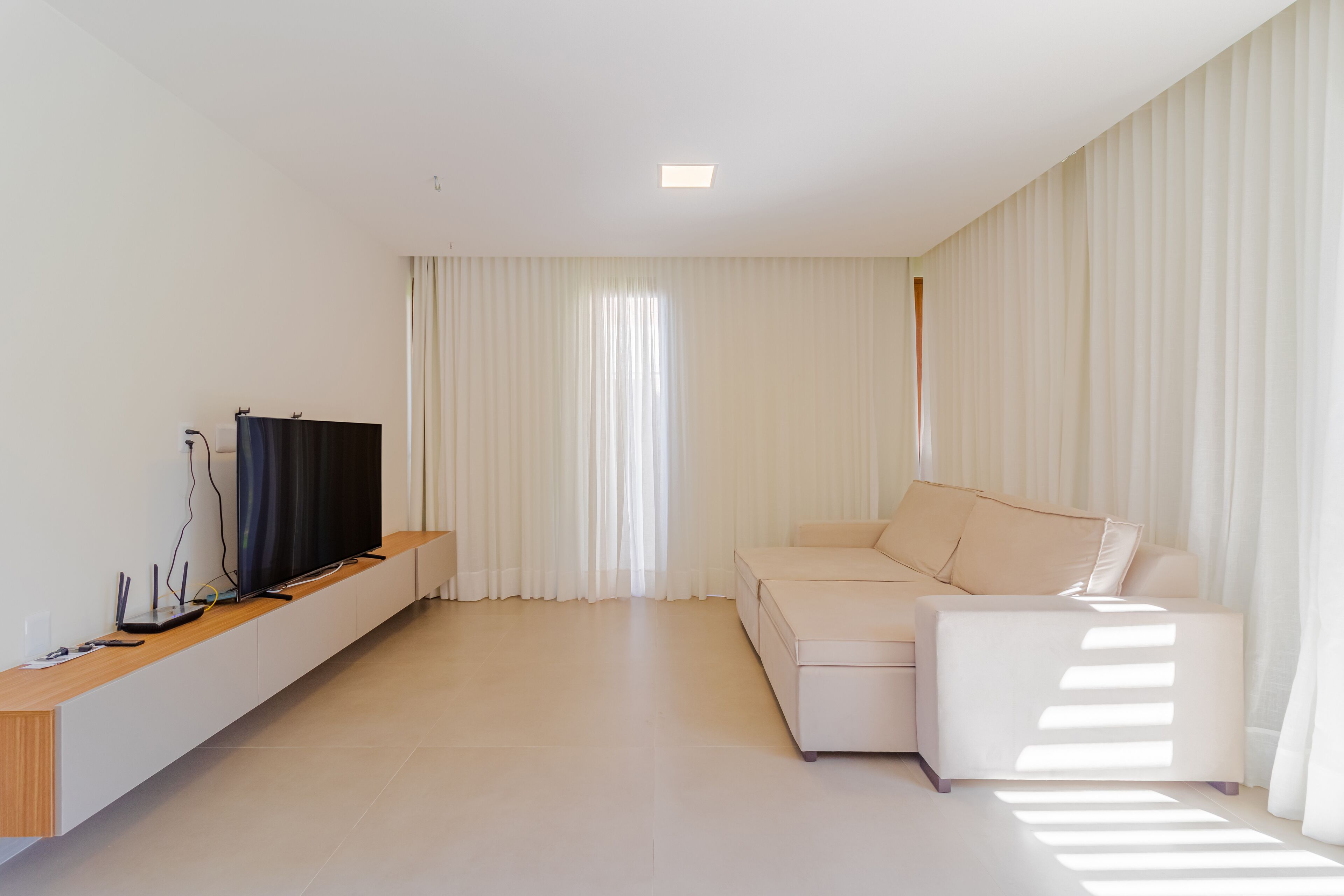 Ein modernes Wohnzimmer mit einem Flachbildfernseher auf einer Holzkonsole, einem beigefarbenen Sofa und einem großen Fenster mit transparenten Vorhängen.