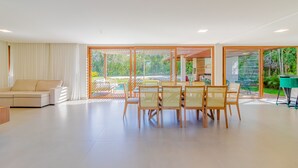 A spacious living area with a sofa, dining table, and chairs. Large windows offer views of greenery and a pool.