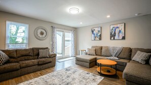 Ein Wohnzimmer mit zwei Sofas, einem Couchtisch und einem Teppich. Zwei Fenster mit Jalousien, eine Tür, die nach draußen führt, und zwei abstrakte Gemälde an der Wand.
