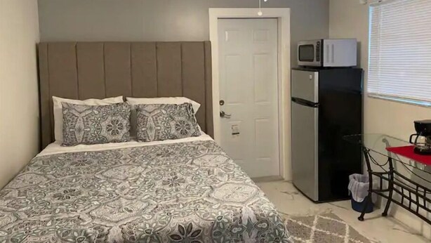 A bedroom with a bed, a ceiling fan, a door, a refrigerator, and a window with blinds. - Private suite, private entrance. (Deerfield Beach)