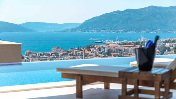 Vista sulla piscina con una panchina di legno e un contenitore nero, che si affaccia su una località costiera e montagne.