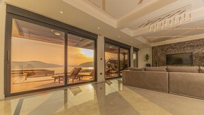 Un'ampia zona giorno con un'ampia porta-finestra scorrevole che conduce a un balcone, un divano moderno, una TV a schermo piatto montata su una parete in pietra e un lampadario.