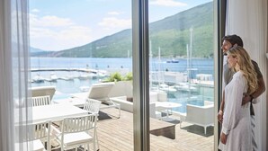 Ein modernes Zimmer mit einem großen Glasfenster, das auf eine Terrasse mit Sitzgelegenheit und Blick auf einen Yachthafen und Hügel führt.