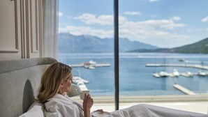 Eine Person in einem weißen Bademantel sitzt auf einem Bett mit Blick auf einen Yachthafen und Berge.