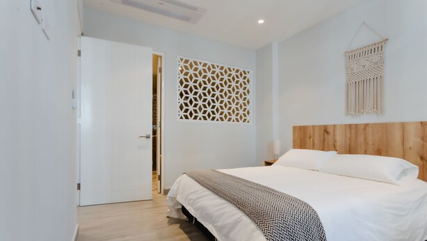 Un dormitorio con cabecera de madera, cama con ropa blanca, manta de diseño y un tapiz de macramé en la pared.