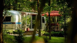 A vintage camper van parked in a forested area with a red canopy and outdoor seating. - Luxury, modern AIRSTREAM at June Farms! (West Sand Lake)