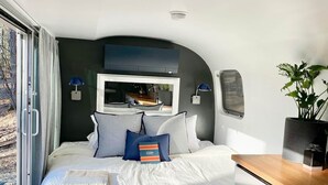 A modern bedroom with a large bed, a built-in desk, and a view through a sliding door. - Luxury, modern AIRSTREAM at June Farms! (West Sand Lake)