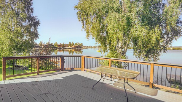 Una terraza de madera con una mesa de vidrio y bancos, con vista a un lago tranquilo, rodeada de árboles.