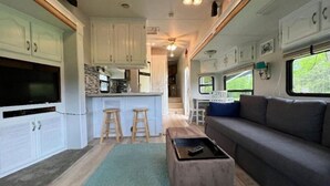 A compact RV interior with a kitchenette, a seating area, and a window providing a view of greenery. - Waterfront at the Pines of Canoe Neck (Abell)