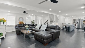 Uma sala de estar moderna com sofá seccional, otomano e mesa de centro. Há um ventilador de teto, iluminação embutida e uma área de cozinha ao fundo.