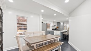 Uma sala de jantar com mesa e bancos de madeira, cozinha ao fundo e vista para uma área residencial pela janela.
