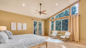 A bedroom with a bed, ceiling fan, large windows, and a view of trees.