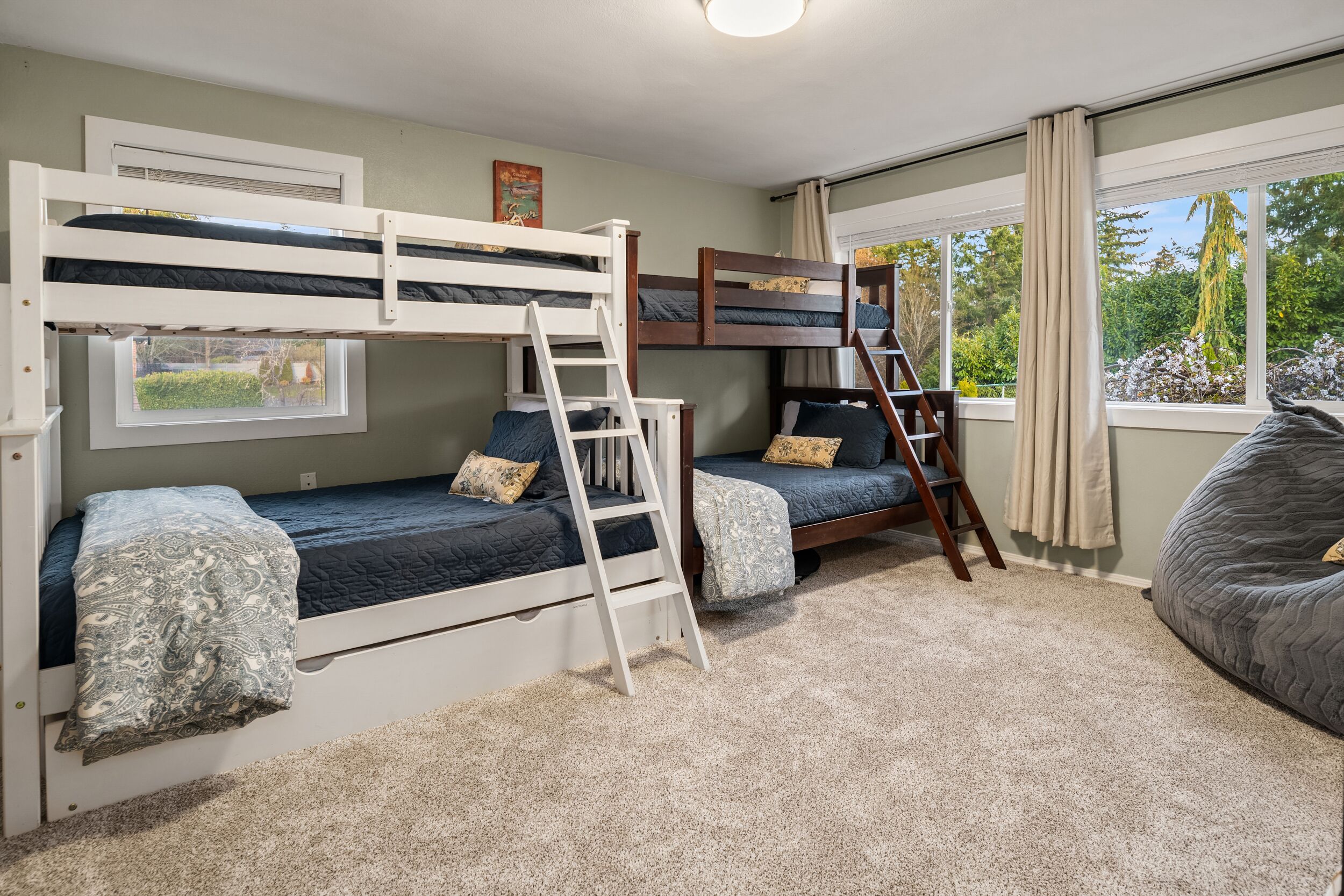 A room with bunk beds, a ladder, a window with a view of greenery, and a bean bag chair.