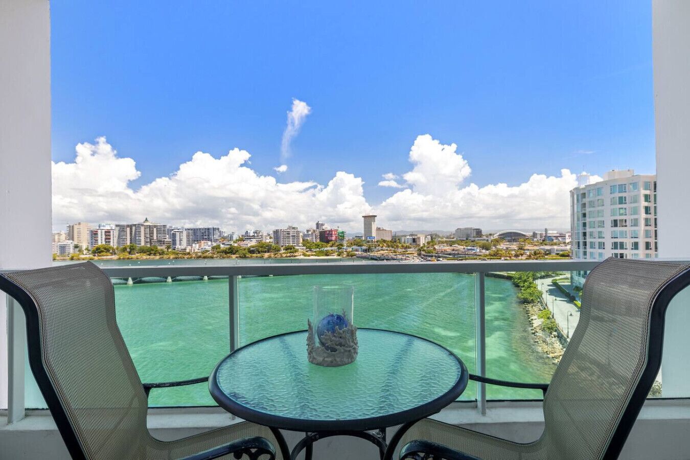 Ein Balkon mit Glastisch, zwei Stühlen und Blick auf eine Stadtlandschaft mit Gebäuden und einer Brücke.