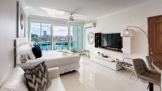 A modern hotel room with a bed, a TV, a dining area, and a balcony with a city view. - Sunrise Studio · Balcony · Water Backup (San Juan)