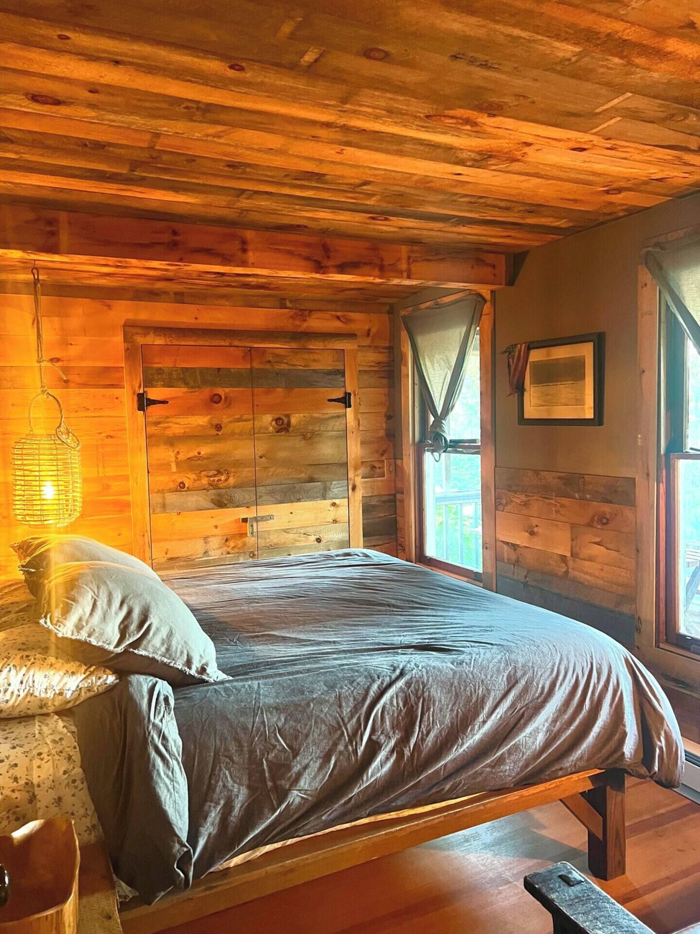 A wooden cabin room with a bed, a hanging light fixture, and a window with curtains.