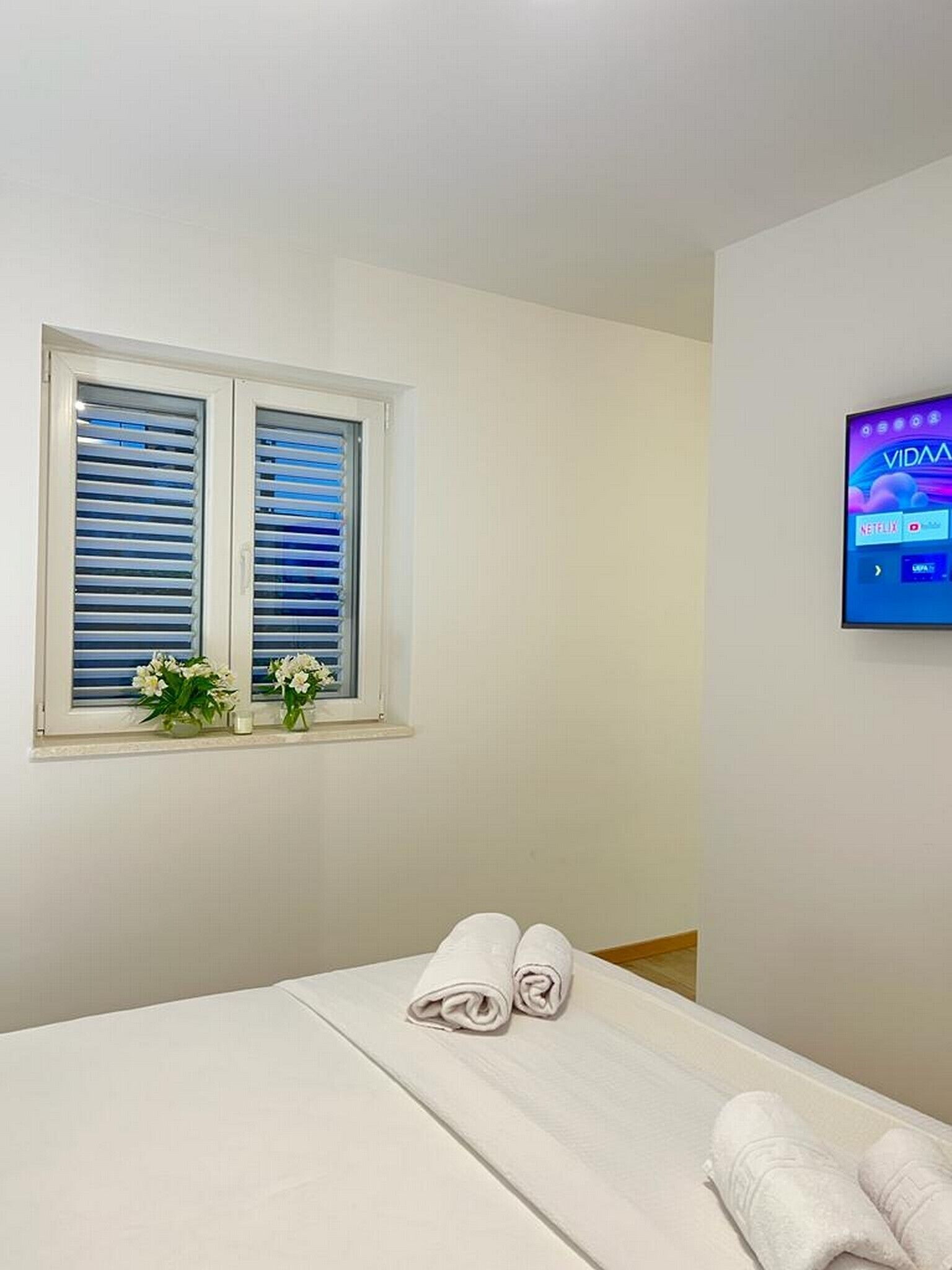 Quarto de hotel com cama, janela com persianas, televisão na parede e uma pequena mesa com um vaso de flores.