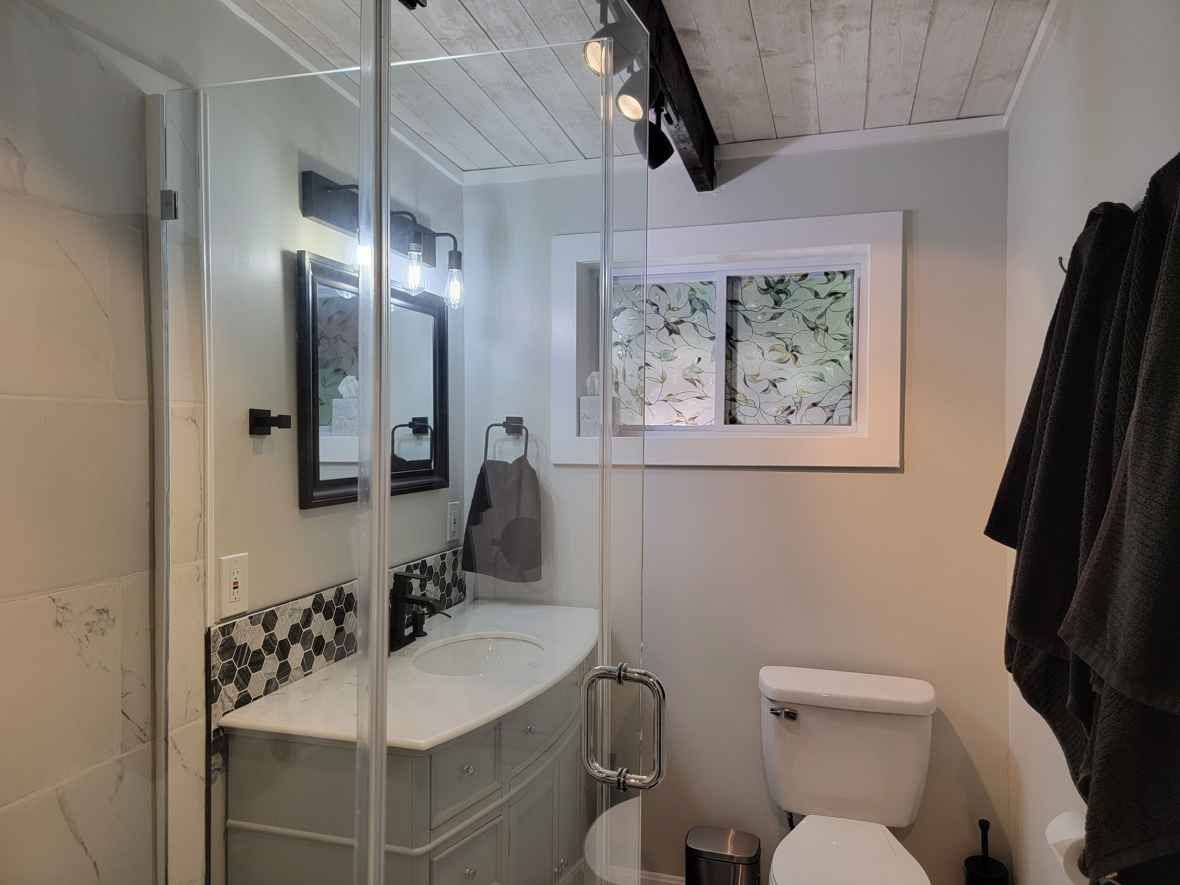 Baño con cabina de ducha de vidrio, lavamanos blanco, inodoro y ventana con estampado floral.