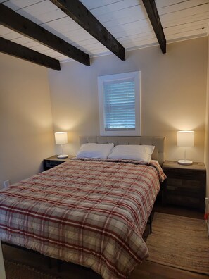 A bedroom with a bed, two bedside tables with lamps, a window with blinds, and exposed wooden beams on the ceiling. - Enjoy Lake life in this renovated Camp, 50 ft from water. Private dock, beach. (Richmond)
