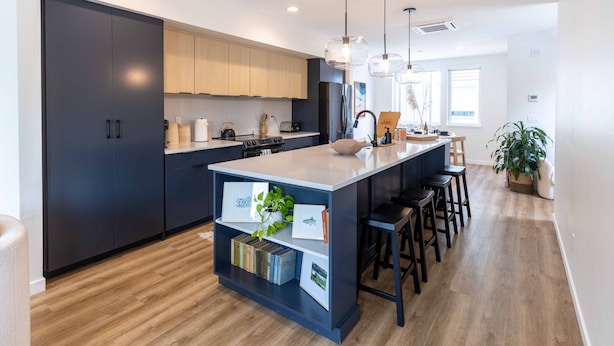 A modern kitchen with dark cabinets, a central island with bar stools, pendant lighting, and a wooden floor.